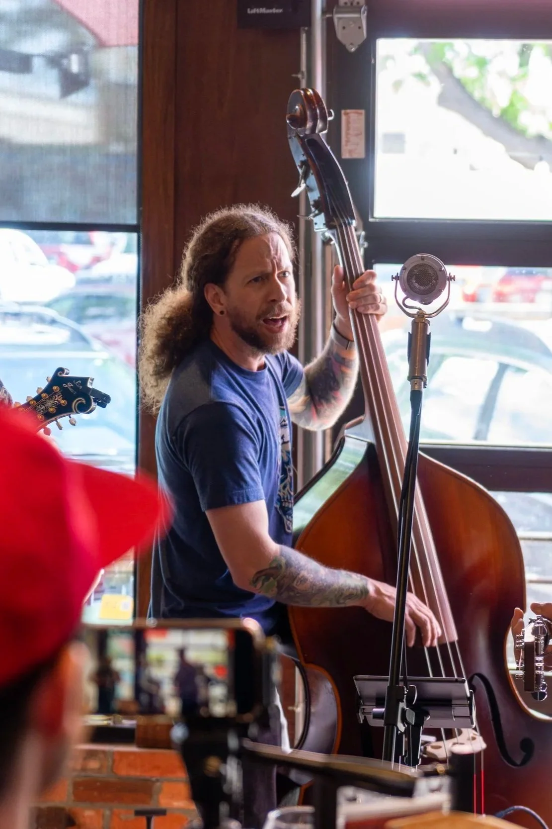A man with long hair and tattoos playing an upright bass in a room with large windows, a microphone, and a guitar partially visible on the left.