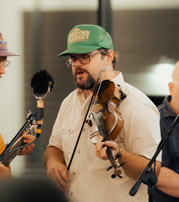 A man with a beard wearing glasses, a green baseball cap, and a beige shirt, playing a violin in a recording studio.