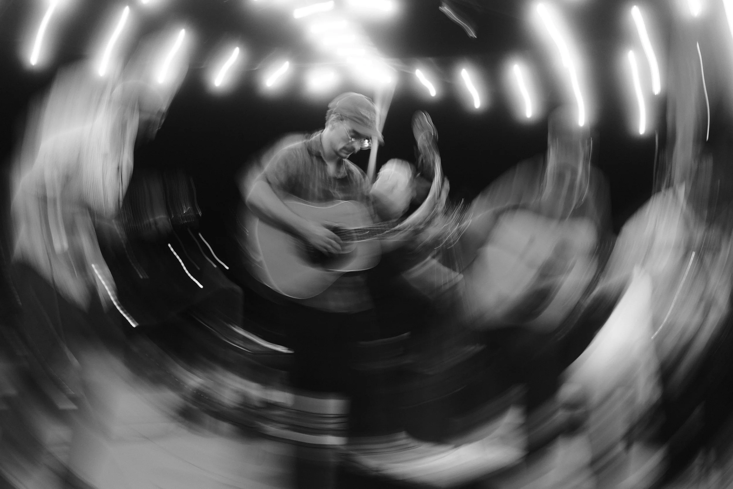 A person wearing a cap and glasses playing an acoustic guitar, with motion blur and lights creating a swirling effect.