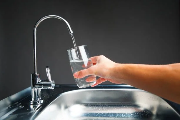 Person filling a clear glass with water from a kitchen faucet over a stainless steel sink.