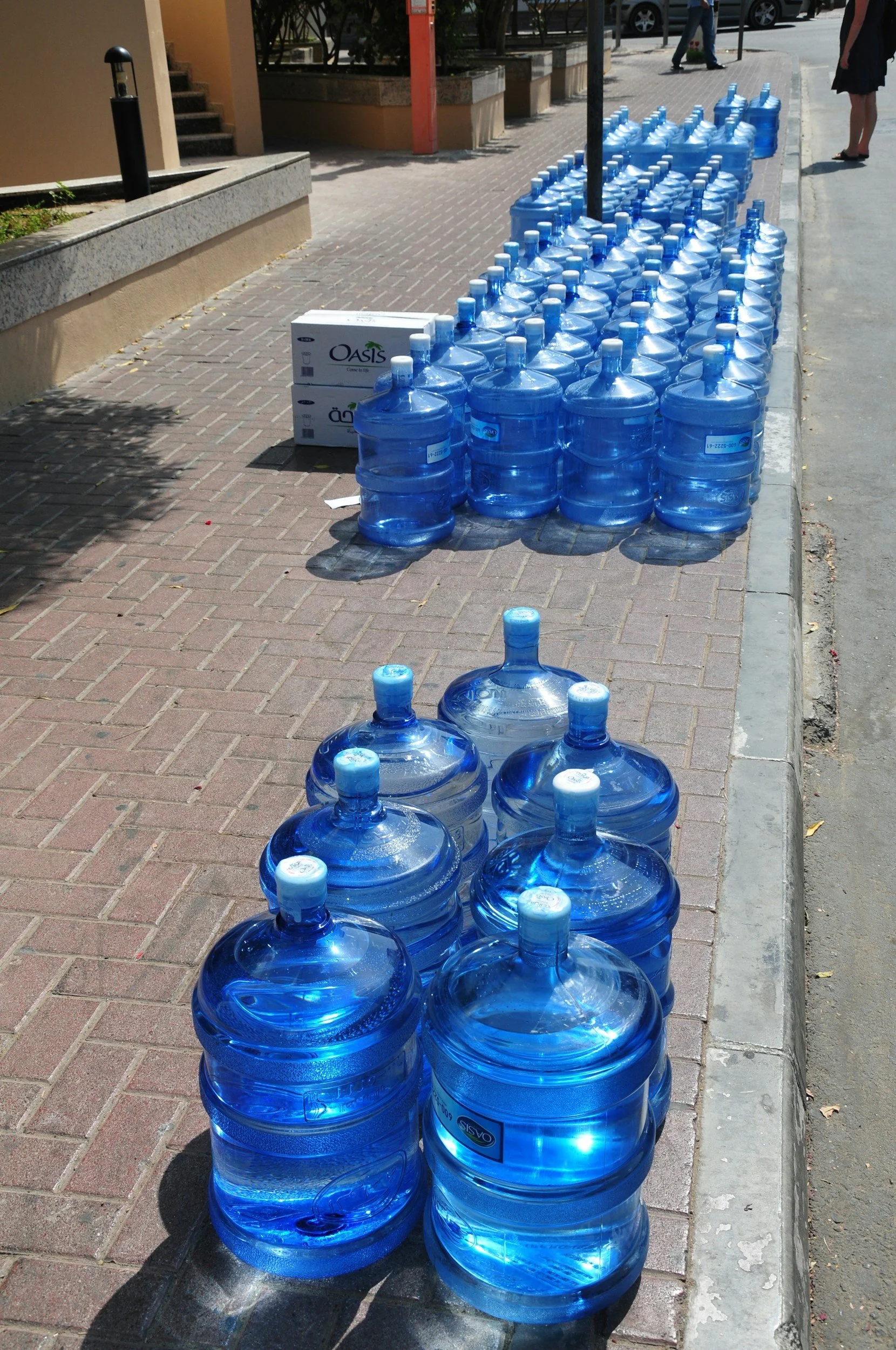 Row of large blue water bottles lined up on a sidewalk with shadows cast on the ground, and a few boxes nearby. A person is walking in the background.