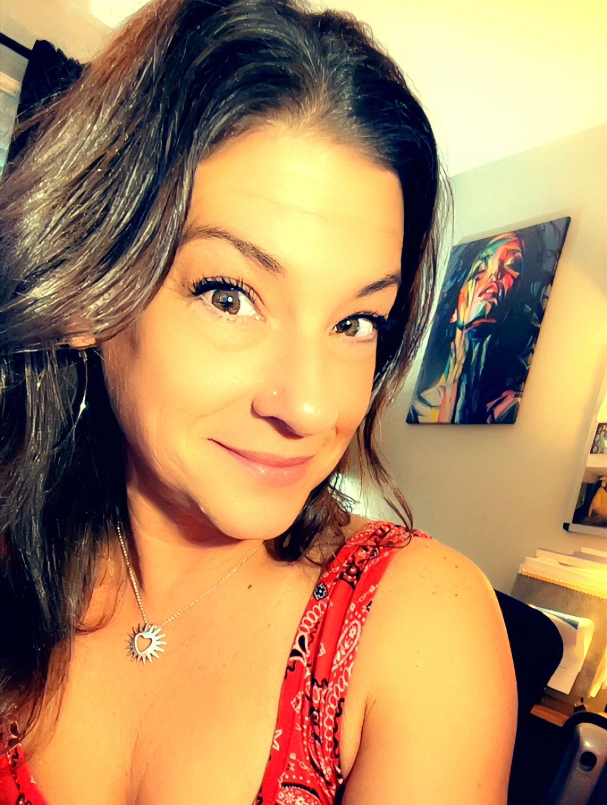 A woman with wavy dark brown hair and brown eyes smiling at the camera, wearing a red patterned sleeveless top and a sun-shaped necklace with a heart in the center. In the background, there is an abstract colorful painting on the wall.