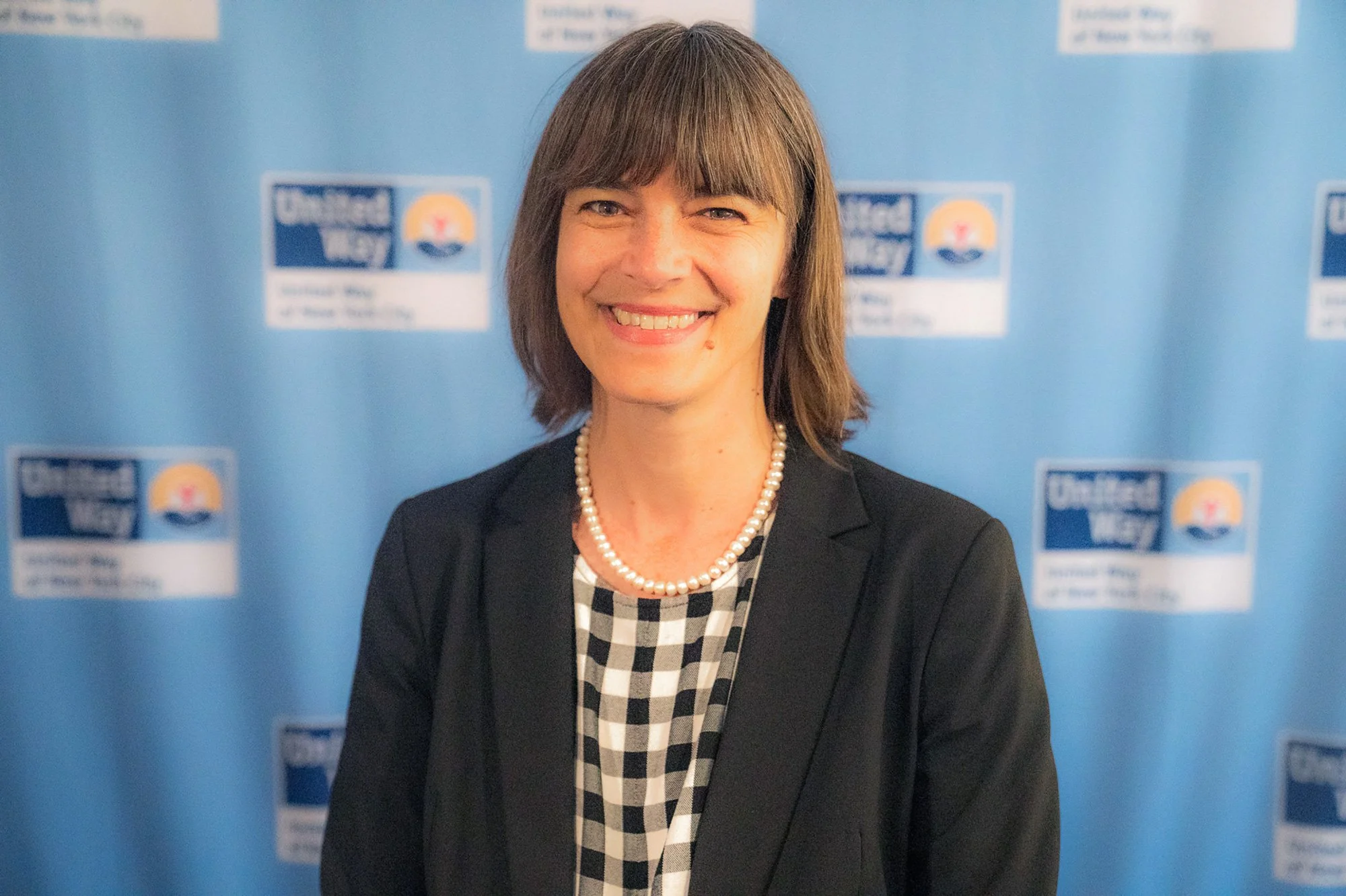 A smiling woman with brown hair, wearing a black blazer, a pearl necklace, and a black-and-white checkered dress, stands in front of a blue backdrop with logos.