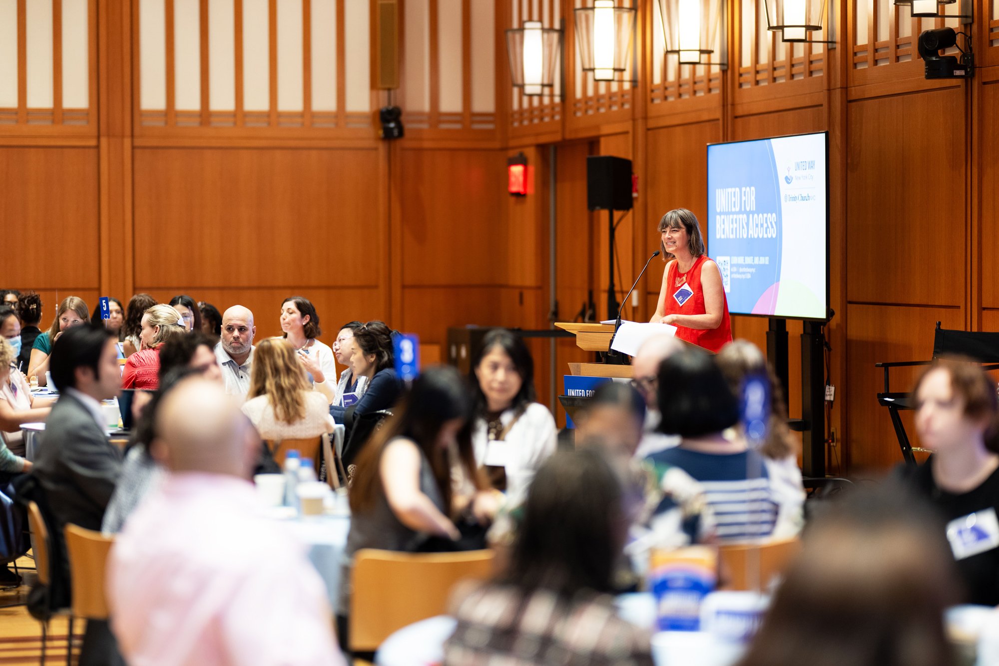 A woman in a red dress speaking at a podium in a large conference room filled with seated attendees. Each side of the room has wooden paneling and lighting fixtures. A large monitor behind the speaker displays the text "United for Benefits Access".