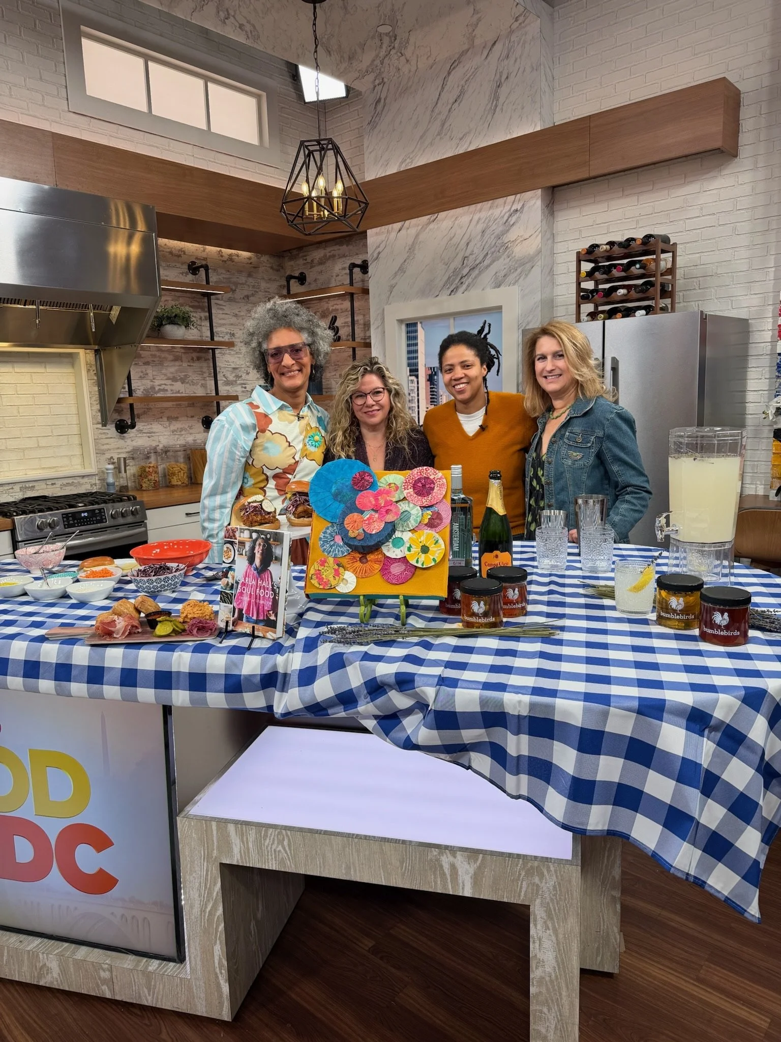 A group of four women standing behind a table with food and drinks, in a modern kitchen with white brick walls, wooden shelves, and a window. The table has a blue and white checkered tablecloth and various food items, jars, and beverages.
