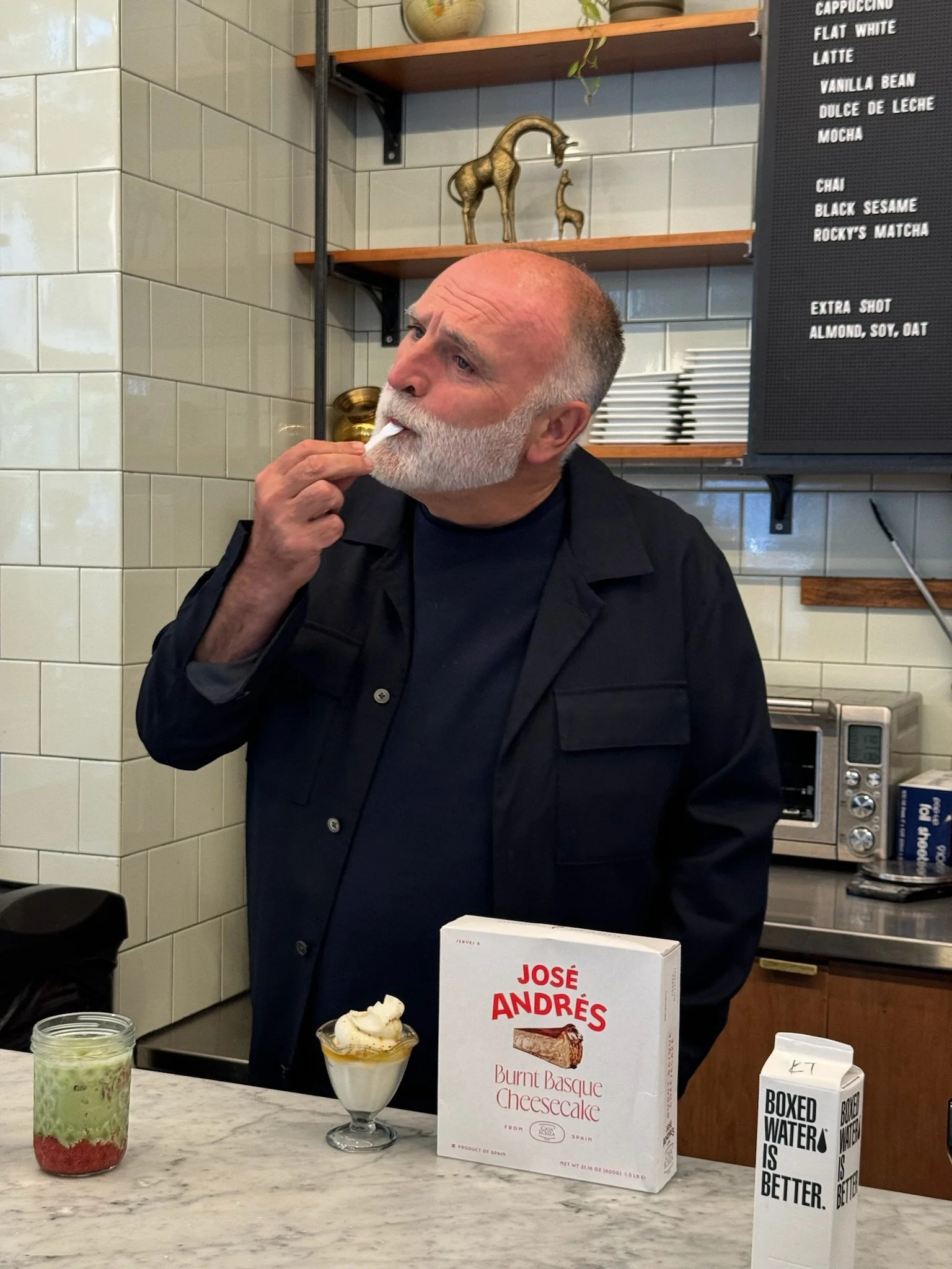 A man with a white beard and bald head is tasting a spoonful of dessert in a café. The decor includes shelves with decorative animal figurines, a menu board listing coffee drinks, and a marble counter with a box of burnt basque cheesecake, a layered beverage, and boxed water on it.