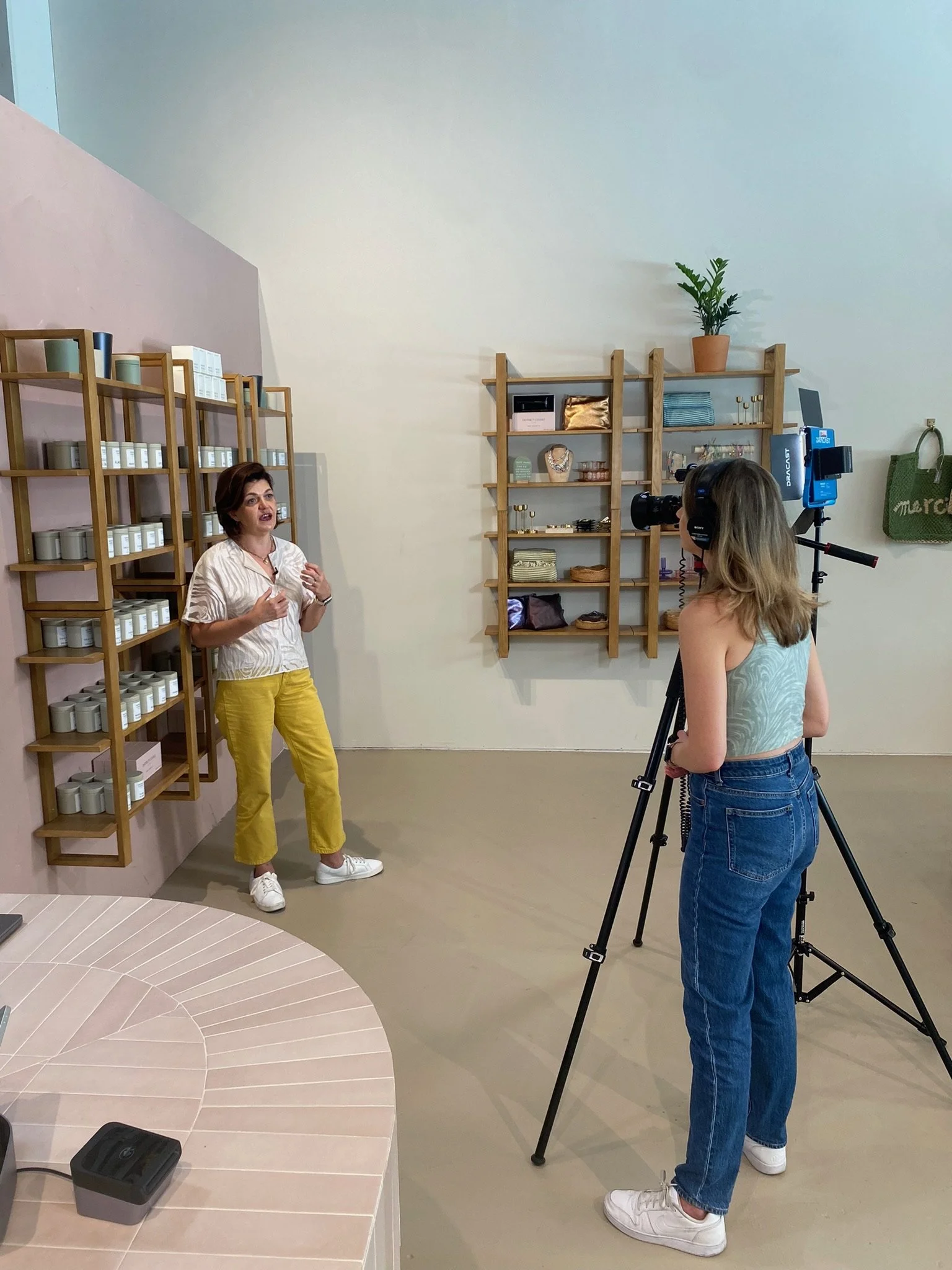 A woman in a white shirt and yellow pants is being filmed by a person with long hair in a tank top and jeans. The woman appears to be giving a presentation or interview in a room with wooden shelves displaying various decorative items and plants.