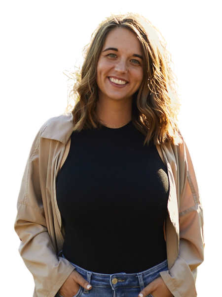 A young woman with shoulder-length wavy brown hair, smiling, wearing a black top and light-colored jacket, standing outdoors.