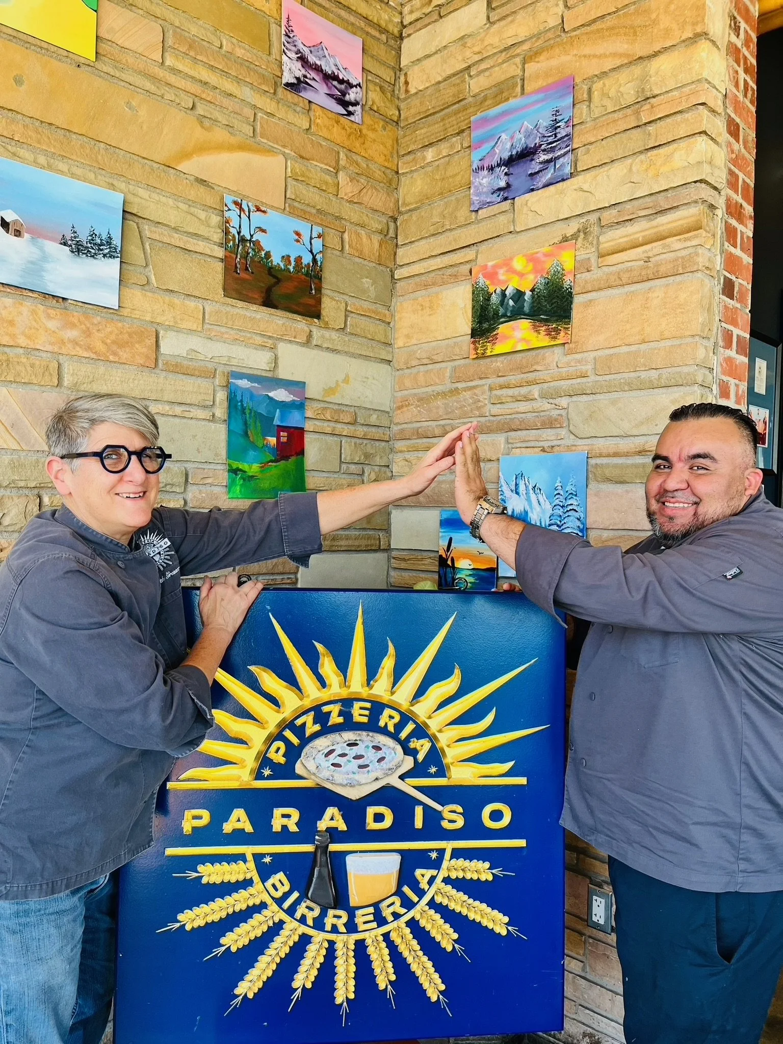 Two people giving a high five in front of a blue sign with yellow text and a logo for a brewery, in a space with a brick wall and several colorful landscape paintings on the wall.