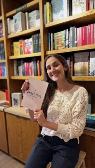 A woman sitting in a bookstore holding a book titled 'Gratitude,' smiling at the camera with bookshelves filled with books behind her.