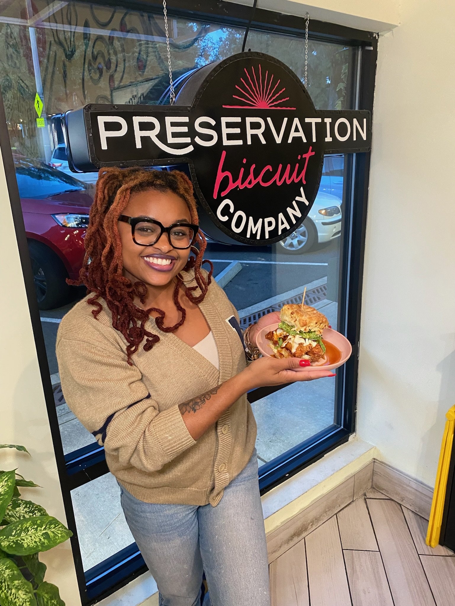 A woman with glasses and dreadlocks smiling and holding a plate with a sandwich inside a restaurant. Behind her is a window with a sign that reads 'Reservation Biscuit Company'.