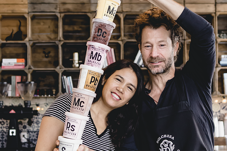 A smiling woman and man make a human tower by stacking gelato containers in a cafe setting. The woman has long dark hair and wears a striped shirt, while the man has curly hair and a beard, wearing a black apron.