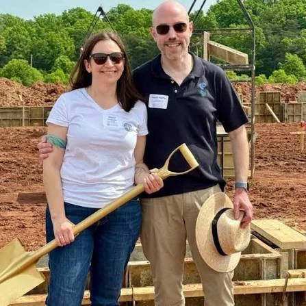 A smiling woman and man at a construction site, holding a shovel and a straw hat, with green trees in the background.