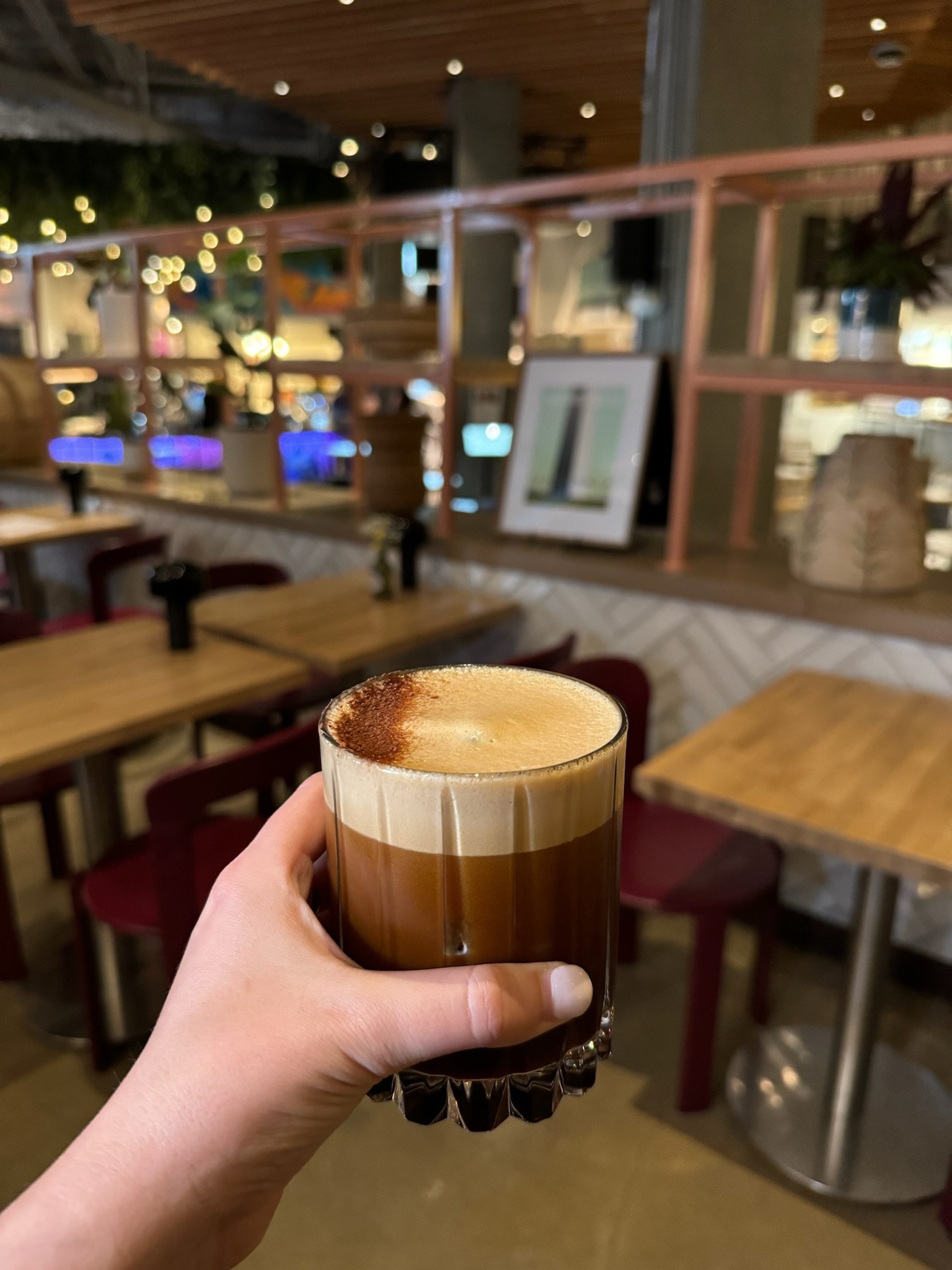 A person holds a glass of layered coffee drink with frothy milk topped with cocoa powder at a restaurant or cafe with wooden tables and chairs.