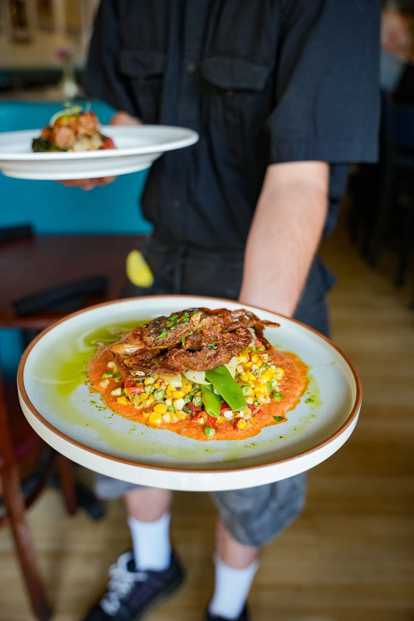 Person holding a plate of colorful food including corn, green beans, tomato-based sauce, and shredded meat topped with herbs, in a restaurant setting.