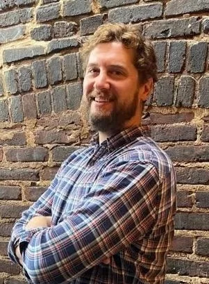 Man with beard and curly hair smiling, wearing a plaid shirt, standing with arms crossed against a brick wall.