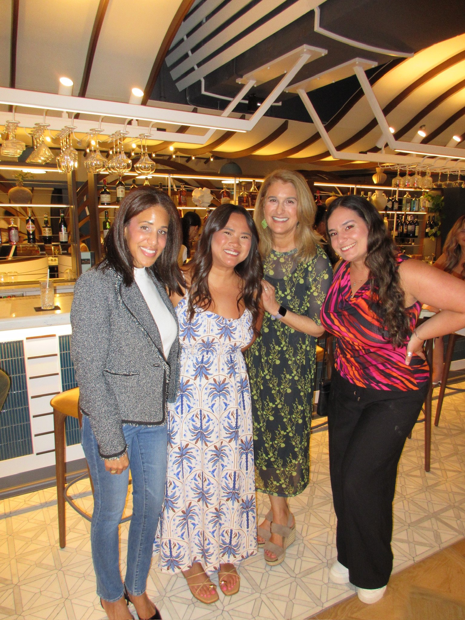 Four women standing together at a bar in a restaurant or bar, smiling at the camera. They are dressed in casual and semi-formal attire, with one wearing a blazer, another in a dress, and the others in tops and pants. The background shows a modern bar setup with bottles, glasses, and hanging wine glasses, under a ceiling with decorative lighting.