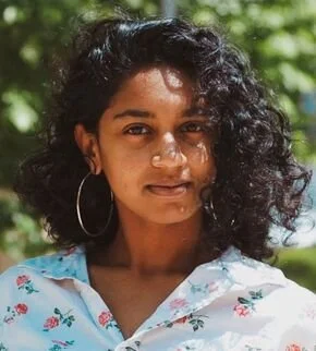 A young woman with curly black hair and hoop earrings, wearing a light-colored floral shirt, standing outdoors with green foliage in the background.