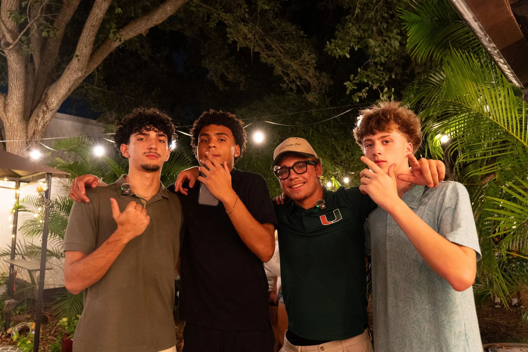 Four young men standing close together outdoors at night, surrounded by trees and string lights, smiling and posing for the photo.