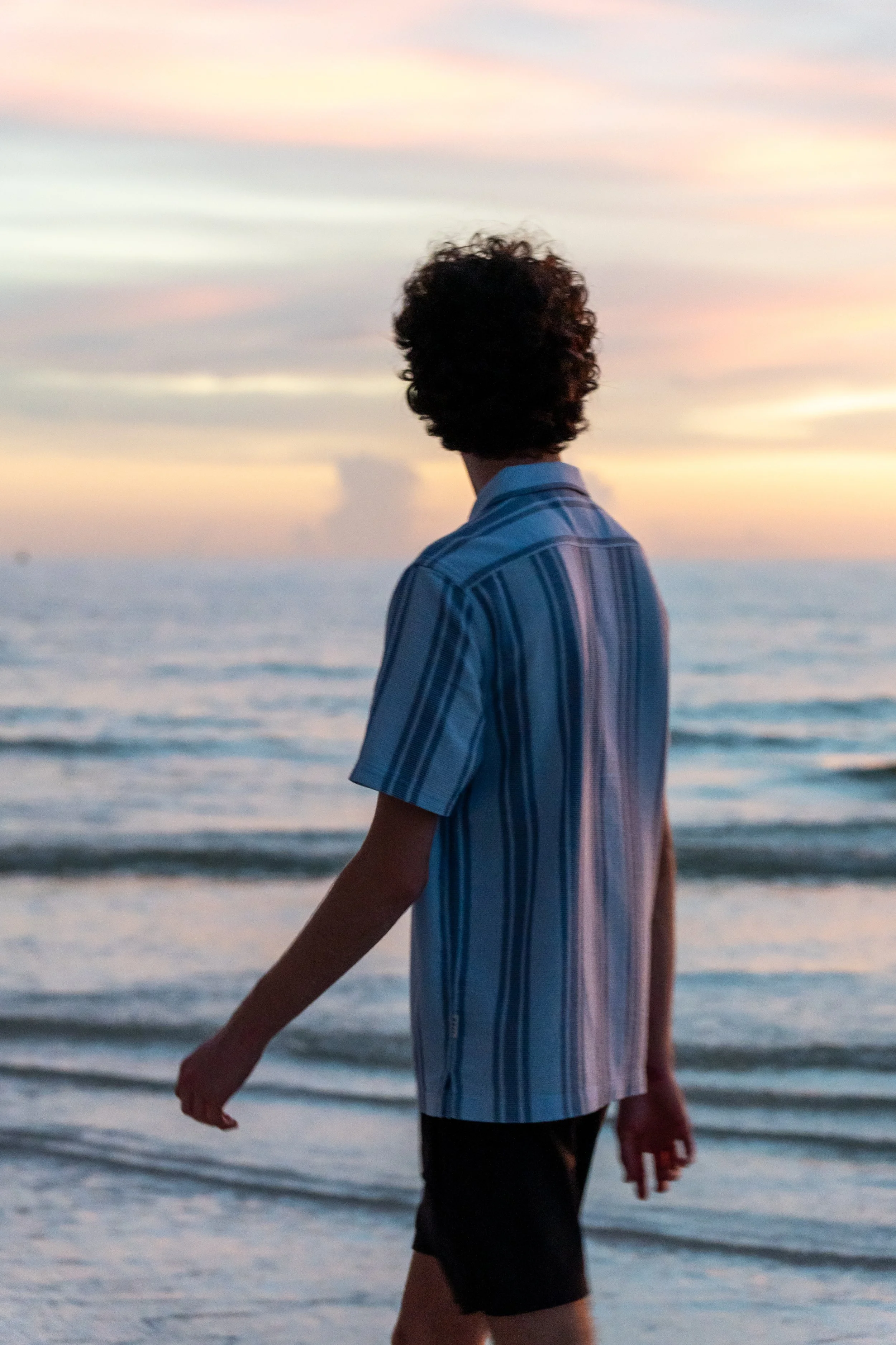 Person with curly hair wearing a striped shirt walking along the beach during sunset.