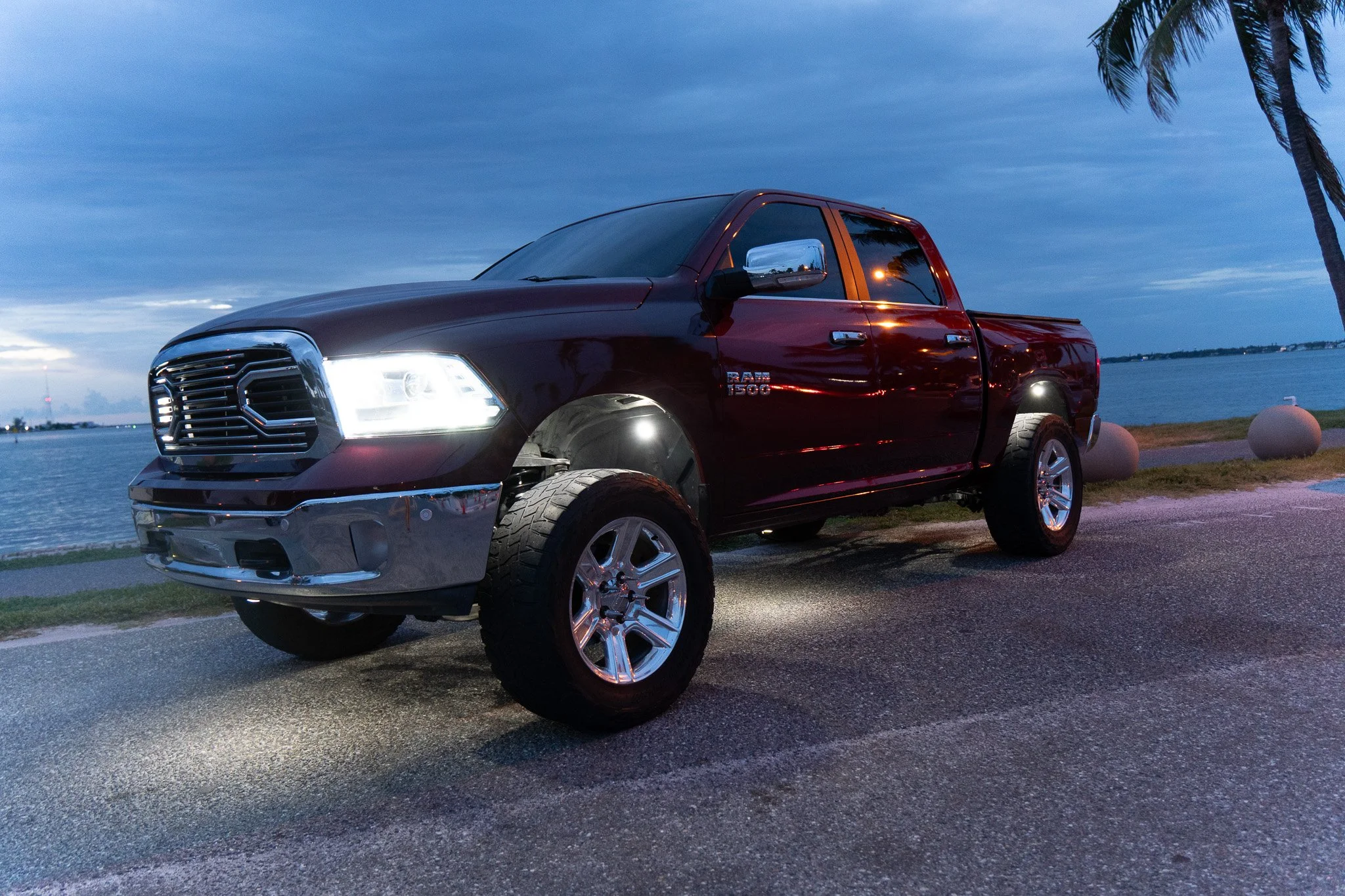 A red RAM 1500 pickup truck parked by the water with palm trees and a cloudy evening sky in the background.