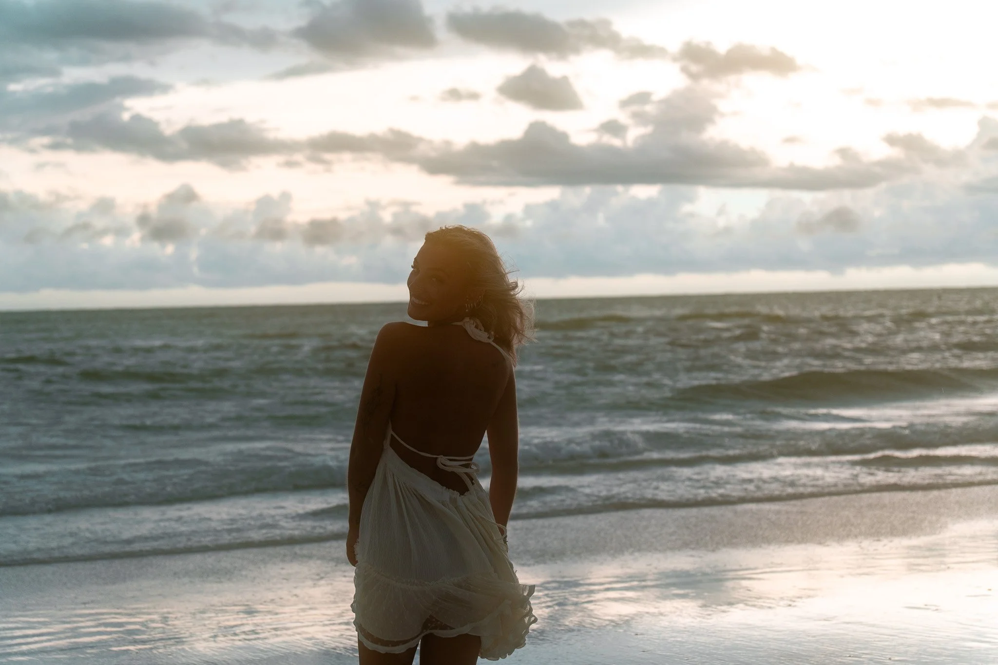 A woman in a light dress standing on the beach looking back over her shoulder with the ocean and cloudy sky in the background.