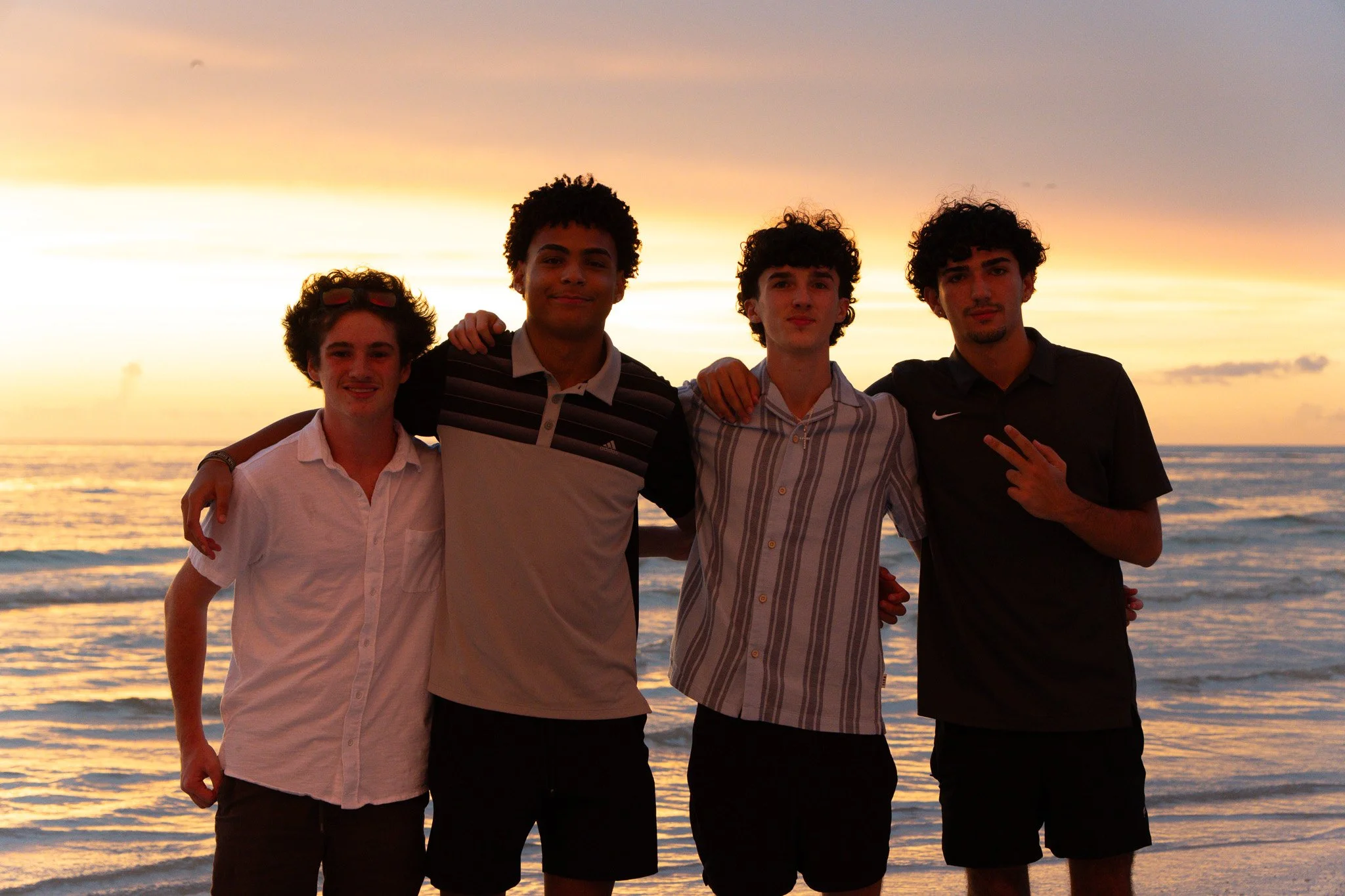 Four young men standing shoulder to shoulder on a beach at sunset, with the ocean in the background, smiling and making hand gestures.