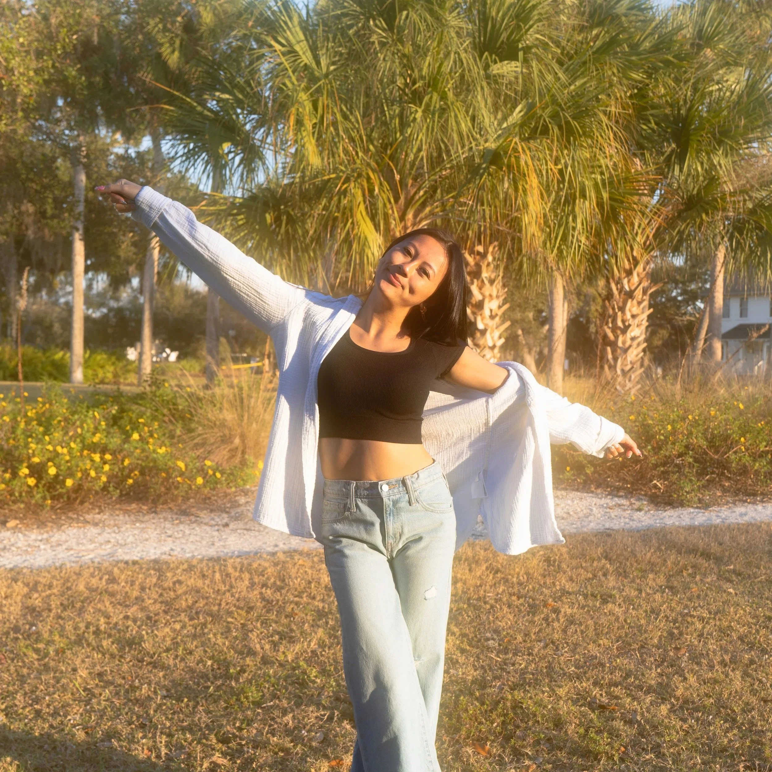 A woman with long dark hair smiling and posing outdoors in front of a large palm tree, with a sunny sky and trees in the background.