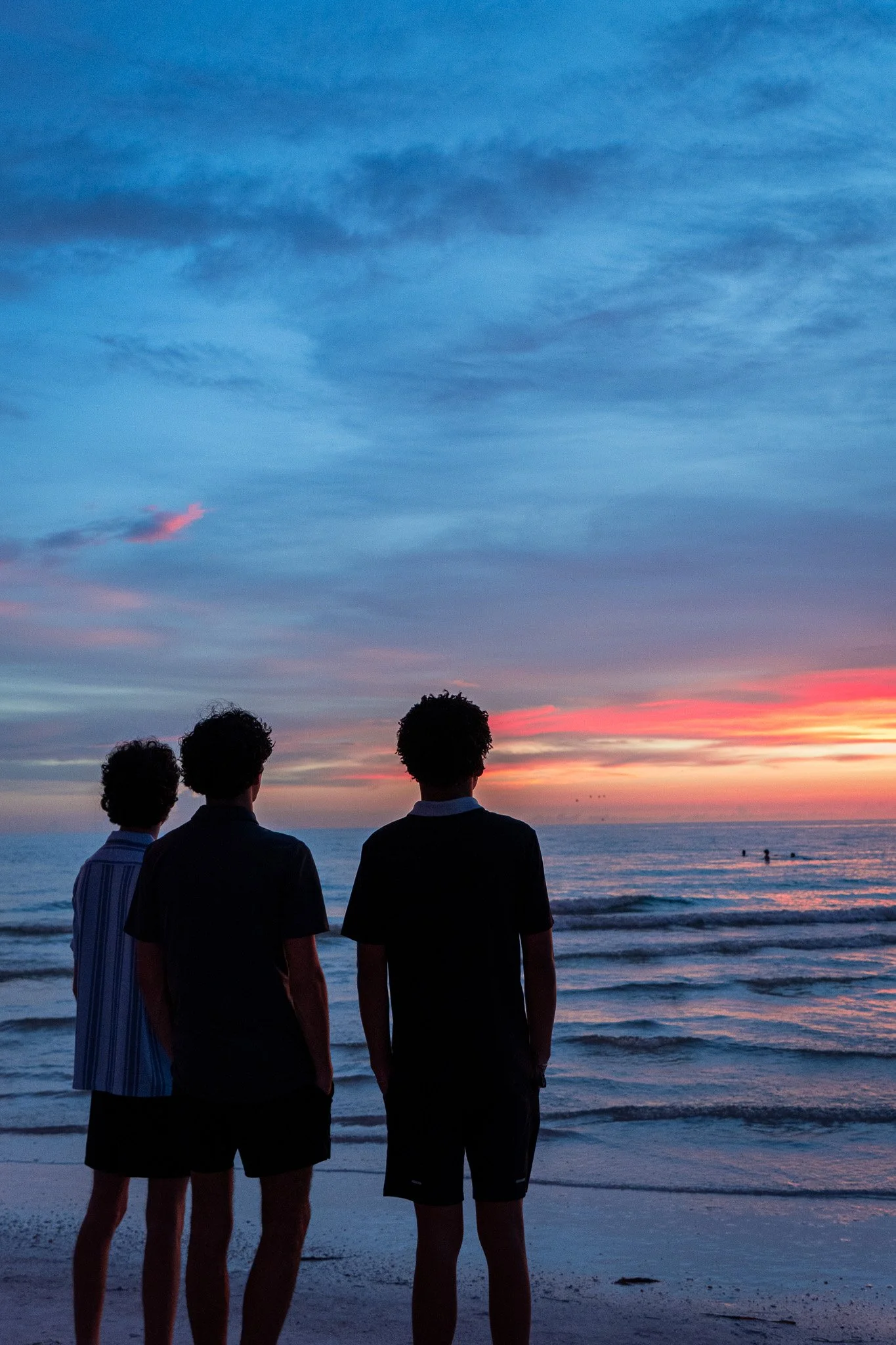 Four people standing on a beach at sunset, facing the ocean, with colorful sky and clouds in the background.