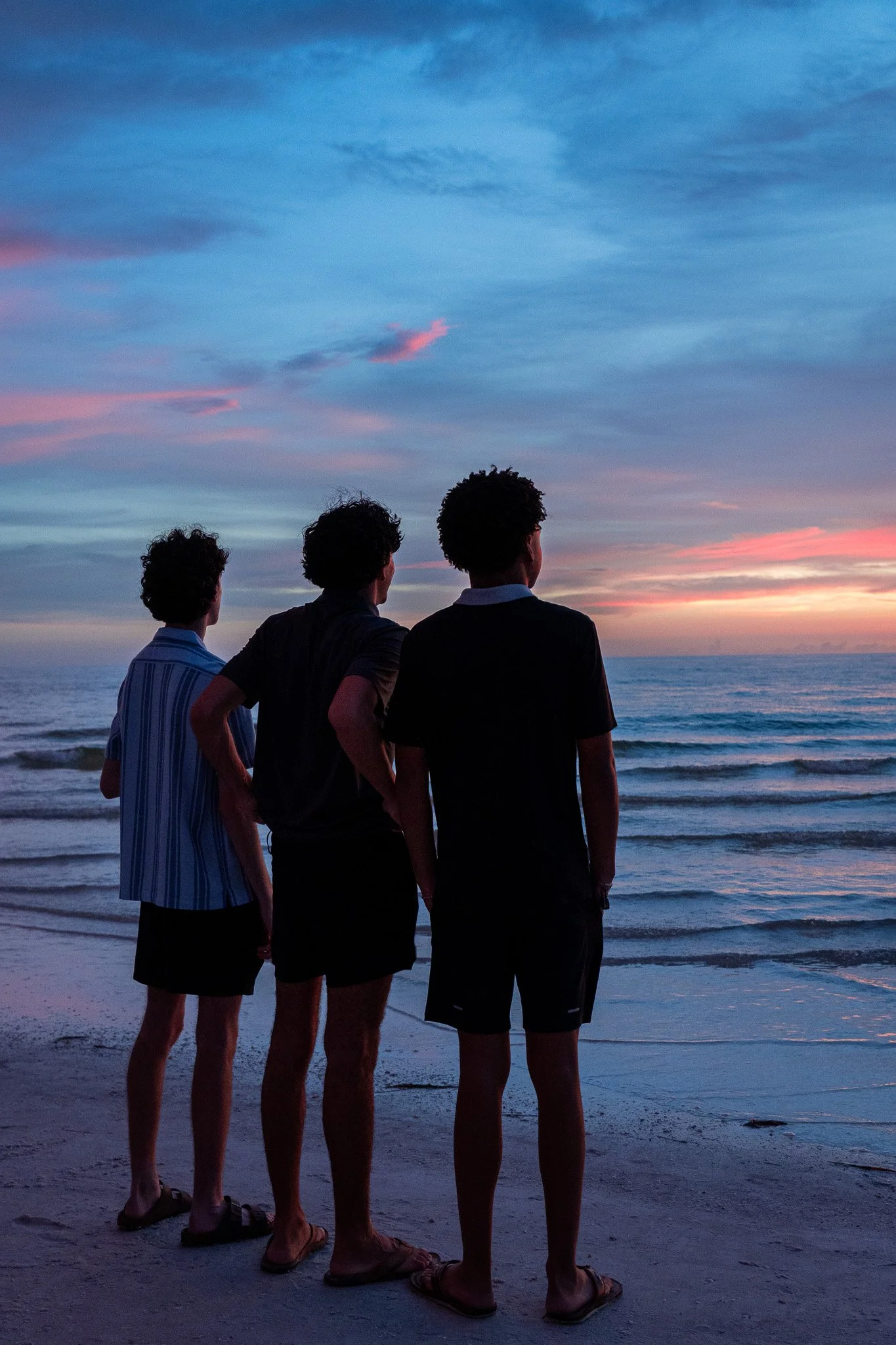 Four young men standing on the beach at sunset, facing the ocean, with clouds in the sky colored pink and blue.