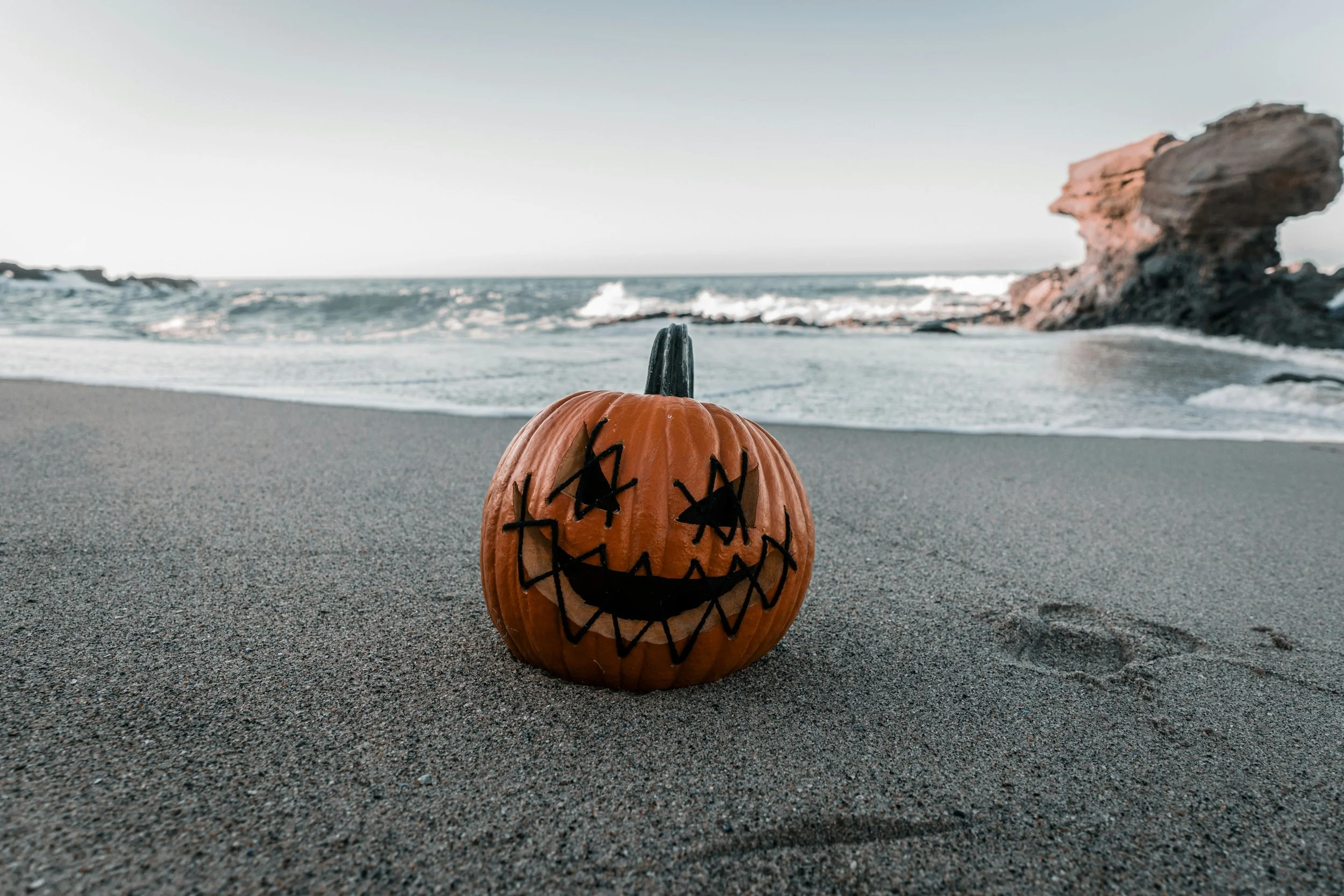 Orange pumpkin with a painted smiling face and eyes, sitting on a sandy beach near the ocean, with a rocky formation in the background.