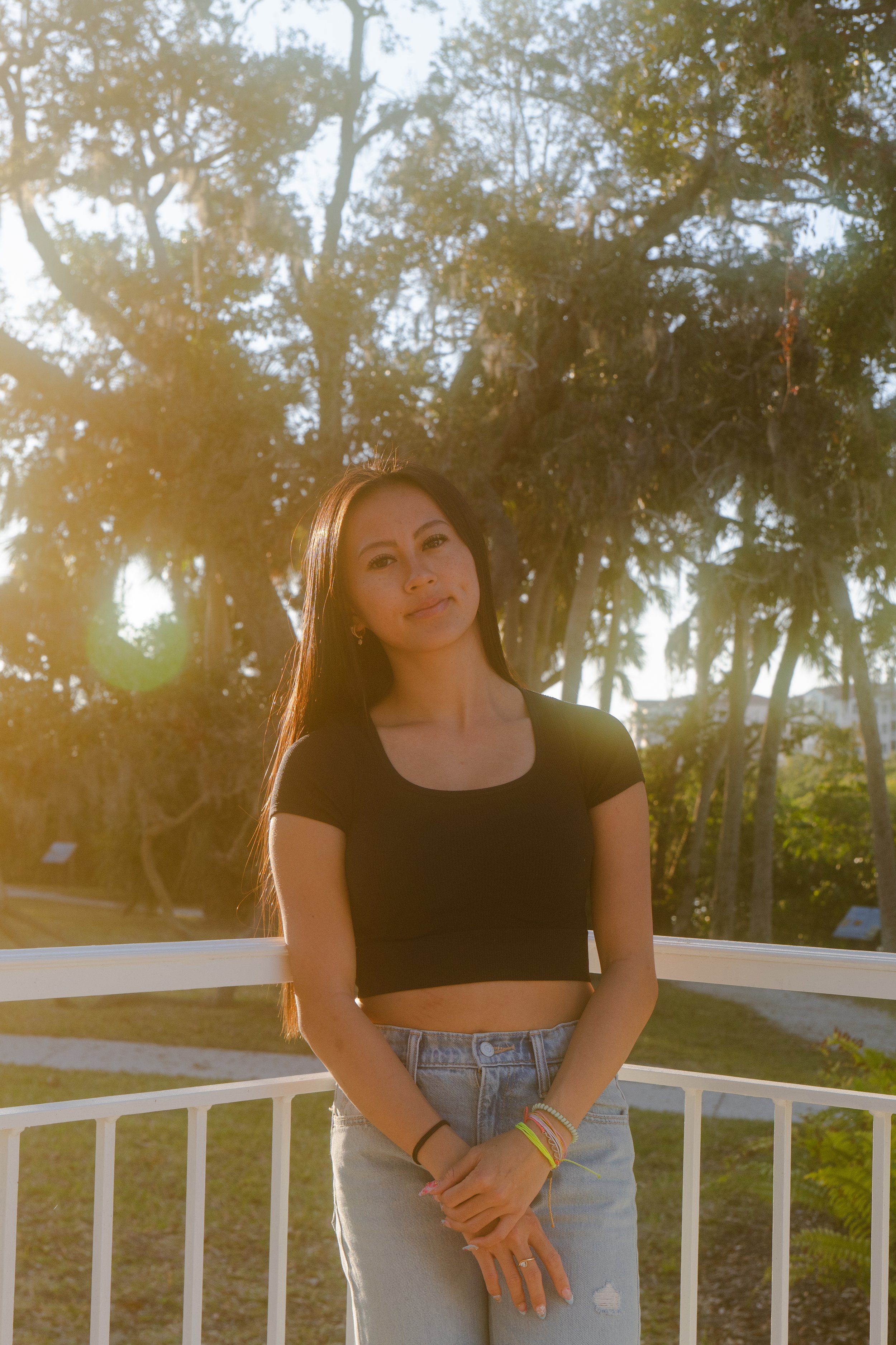 A young woman with long dark hair stands outdoors in front of a white fence, smiling slightly. She is wearing a black crop top, ripped high-waisted jeans, and colorful wristbands. The background features tall trees and sunlight filtering through the leaves, creating a warm and sunny atmosphere.