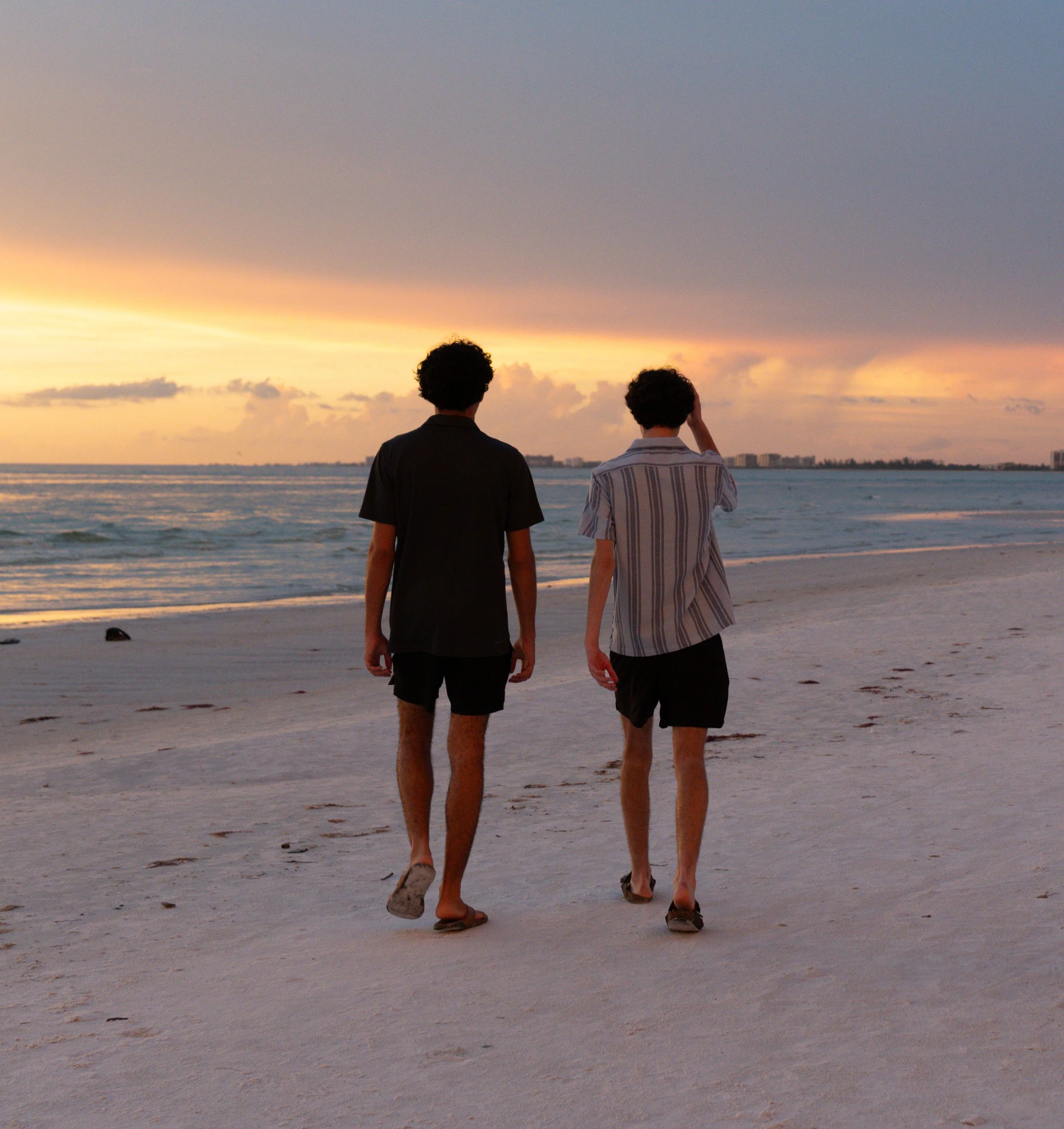 Two young men walking along a beach at sunset, with the ocean and skyline in the background.