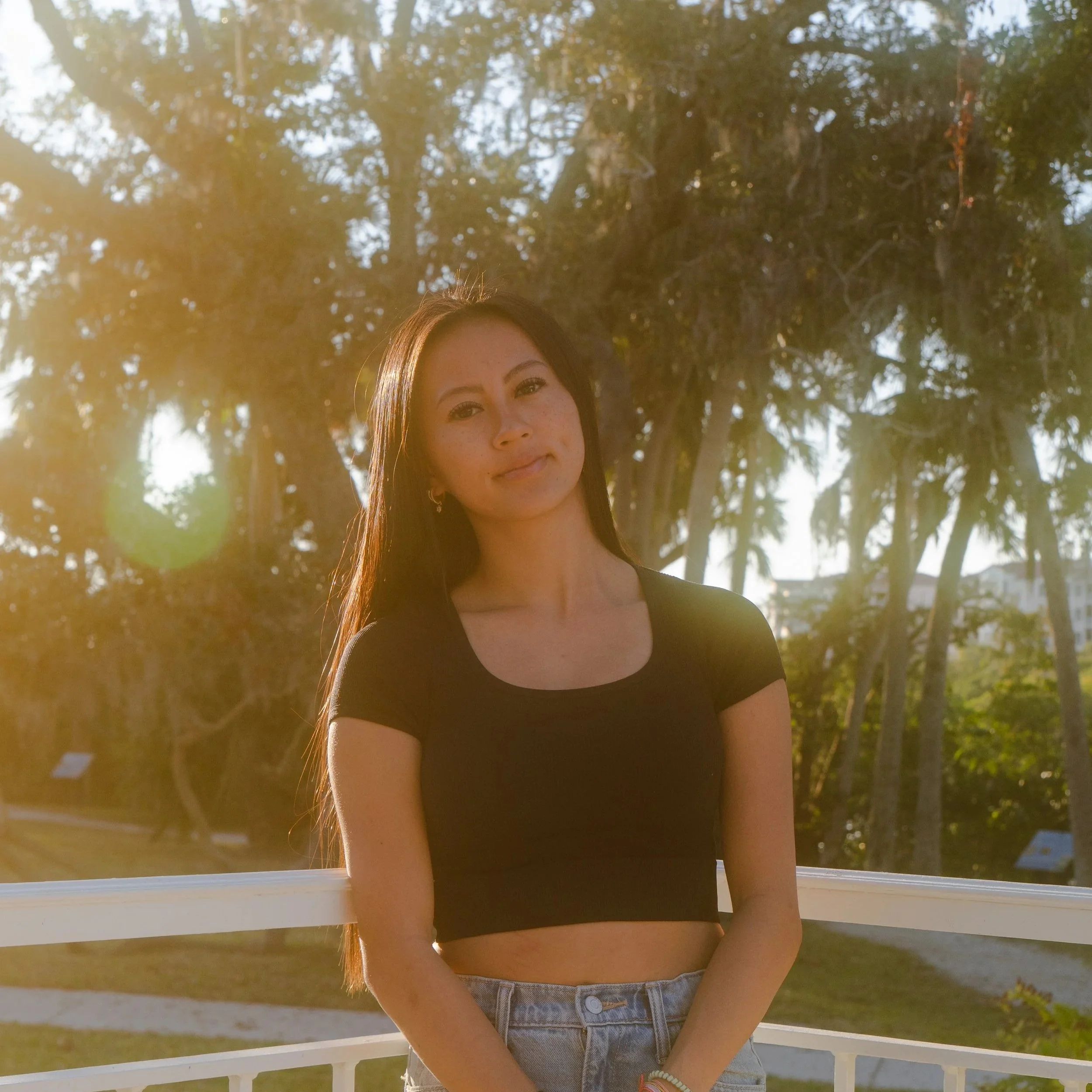 A young woman with long brown hair, wearing a black top and light-colored shorts, standing outdoors near a white fence with trees and sunlight in the background.