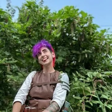Person with purple and pink hair smiling outdoors with green foliage in the background.
