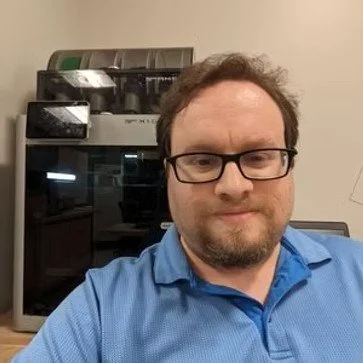 A man wearing glasses and a blue collared shirt taking a selfie in an indoor setting, with a machine or appliance in the background.