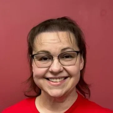 A woman with glasses, short dark hair, and a red shirt smiling in front of a pink background.