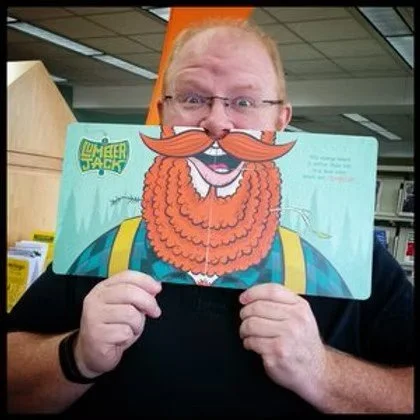 Person holding a Dr. Seuss-style illustration with a large orange beard and mustache, hiding part of his face, in a library or bookstore.