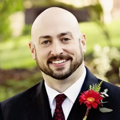 A man with a shaved head and beard, wearing a tuxedo with a red flower boutonniere, smiling outdoors with greenery in the background.