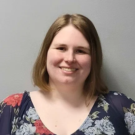 A woman with shoulder-length light brown hair smiling, wearing a floral blouse with navy and red flowers, standing against a plain gray wall.