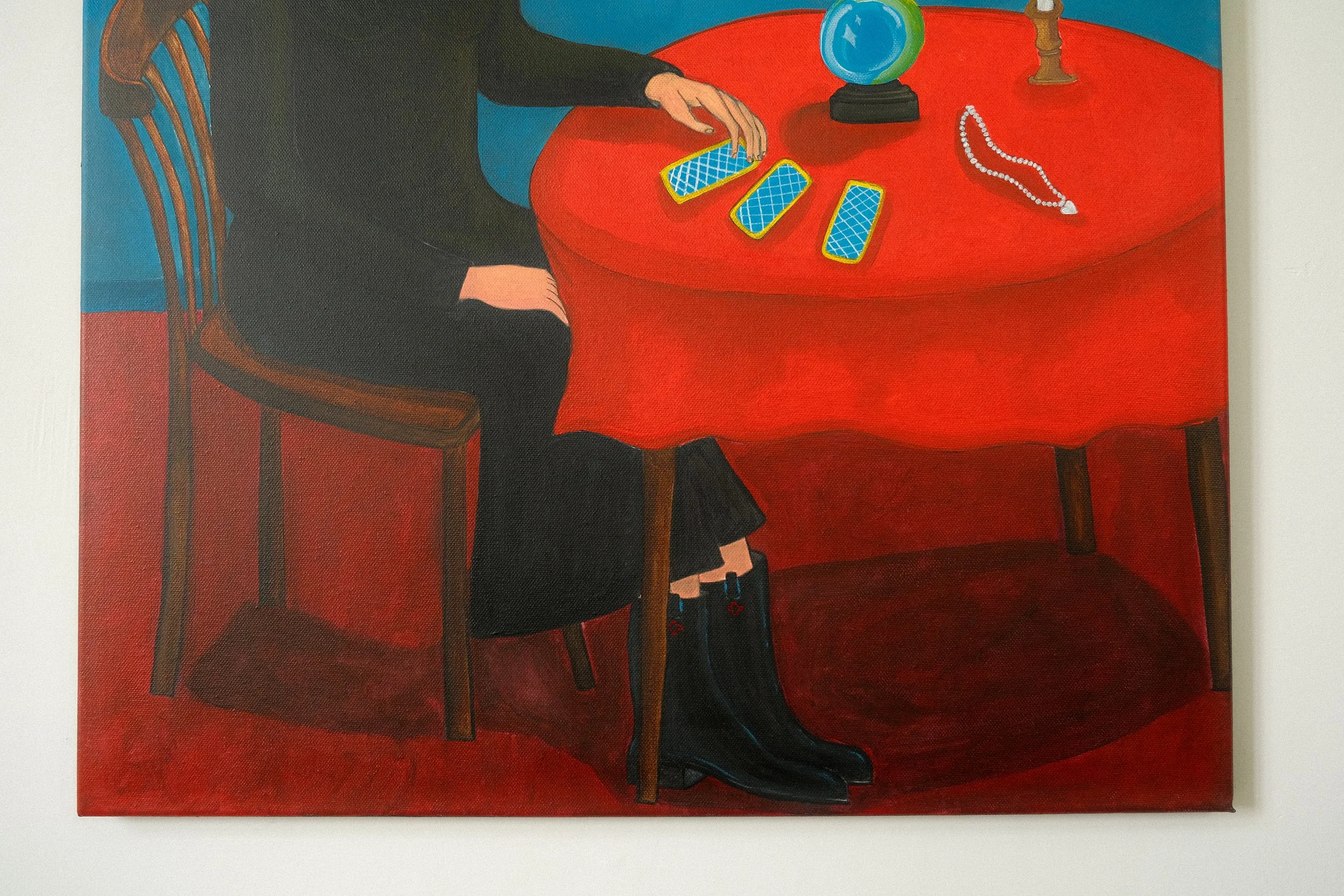 Detail of the painting The Fortune teller. The fortune teller is wearing black clothing, and the scene has a bold, stylized artistic style. There are tarot cards, a candle; a silver necklace and a crystal ball on the table. She wears black boots.