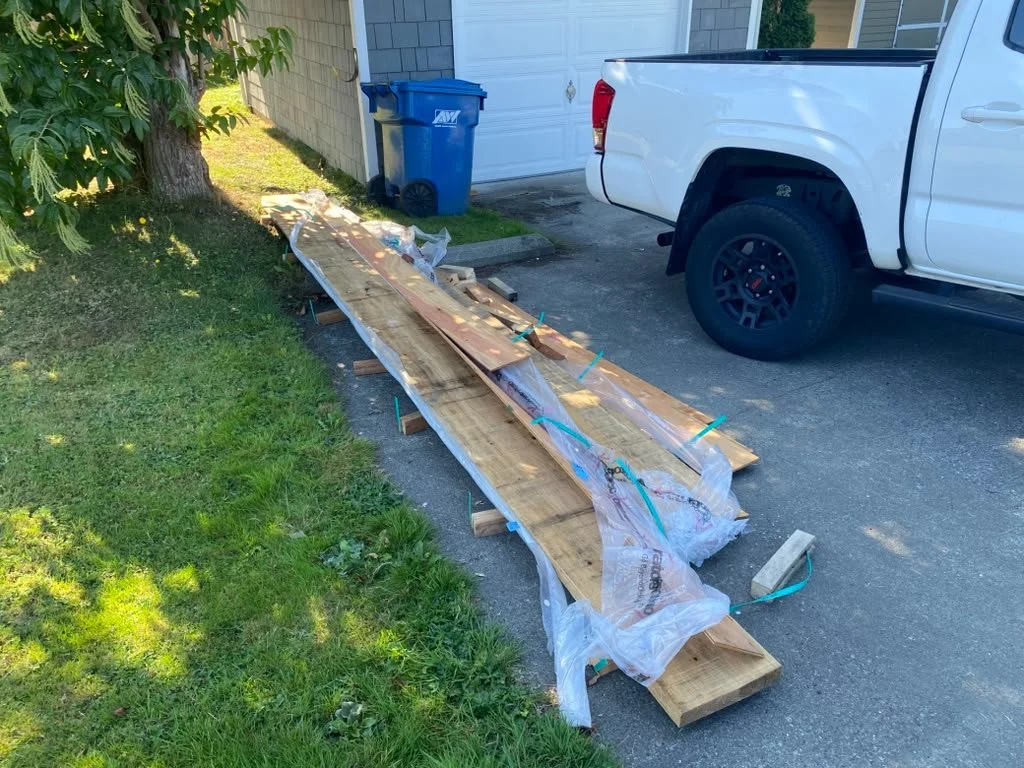 Long wooden boards and planks placed on wooden supports outside near a white pickup truck, with a blue trash bin and a garage in the background.