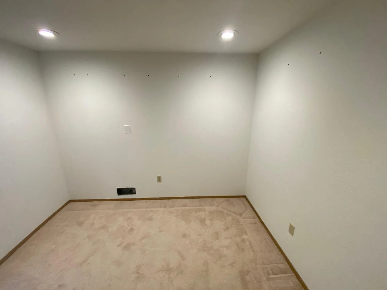 Empty room with white walls, beige carpet, ceiling lights, and electrical outlets.