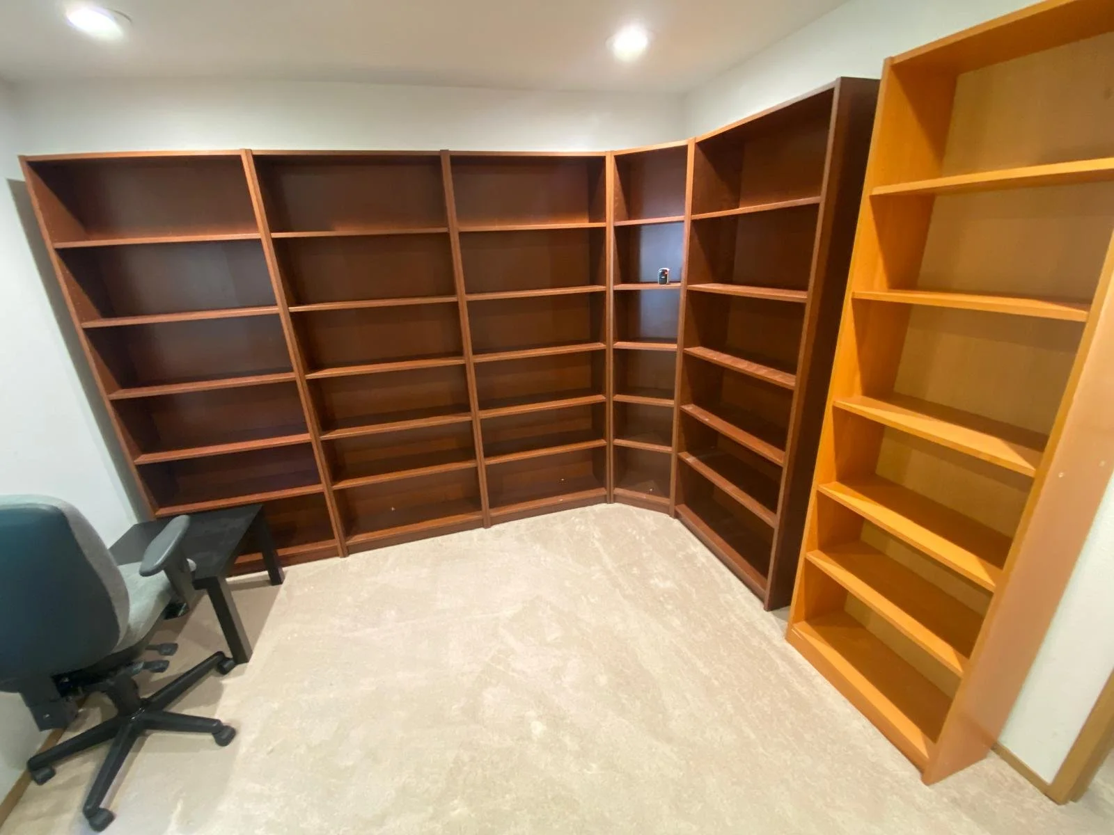 Empty wooden bookshelves in a room with a beige floor, a gray chair, and a small black table.