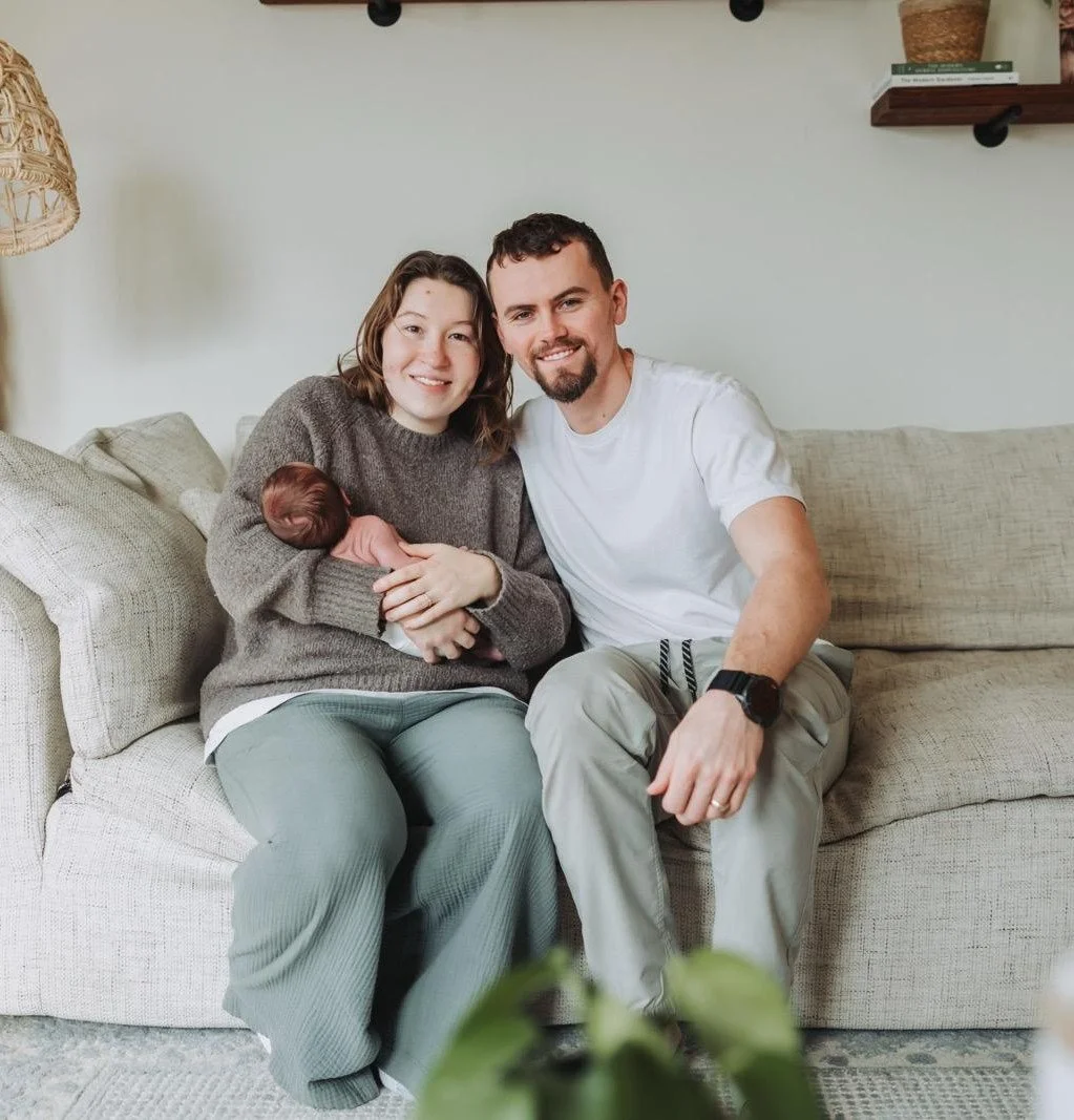 A young woman holding a newborn baby sitting on a gray couch with a young man, all smiling indoors.