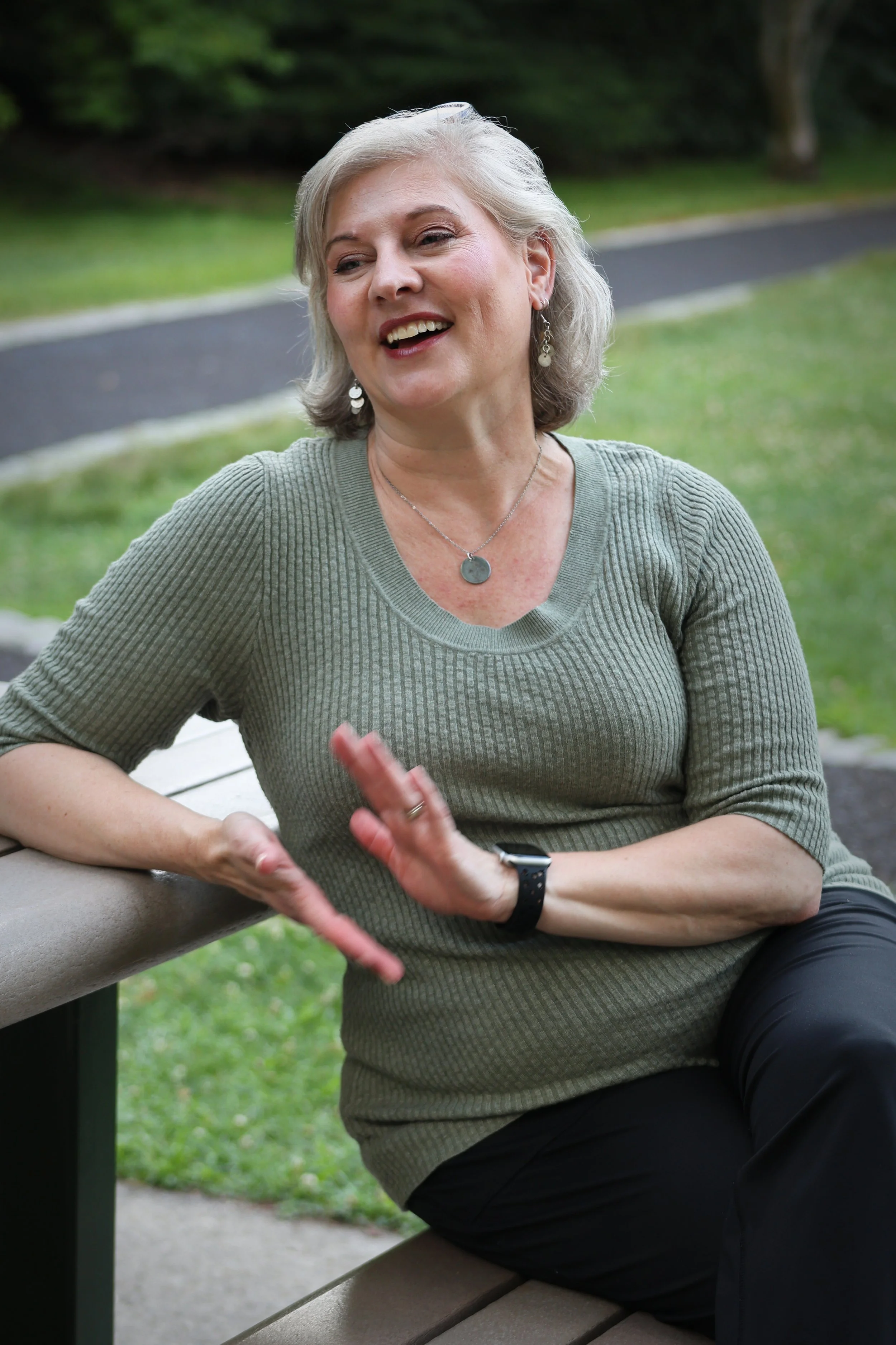 A woman with a friendly expression sitting outdoors by a bench on a grassy area, wearing a green sweater, earrings, a necklace, and a smartwatch, with trees and a pathway in the background.