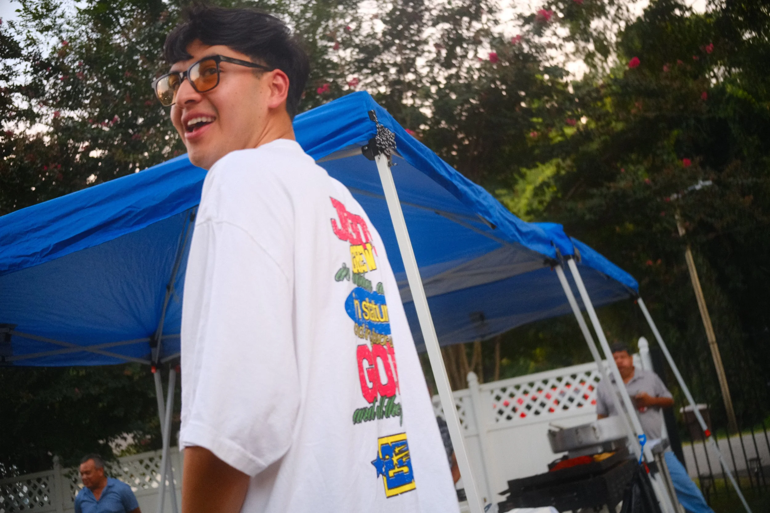 A young man with dark hair, glasses, and a white T-shirt smiling and looking to his right outdoors during the daytime. There is a blue canopy tent behind him, and other people in the background, with trees and a white lattice fence.
