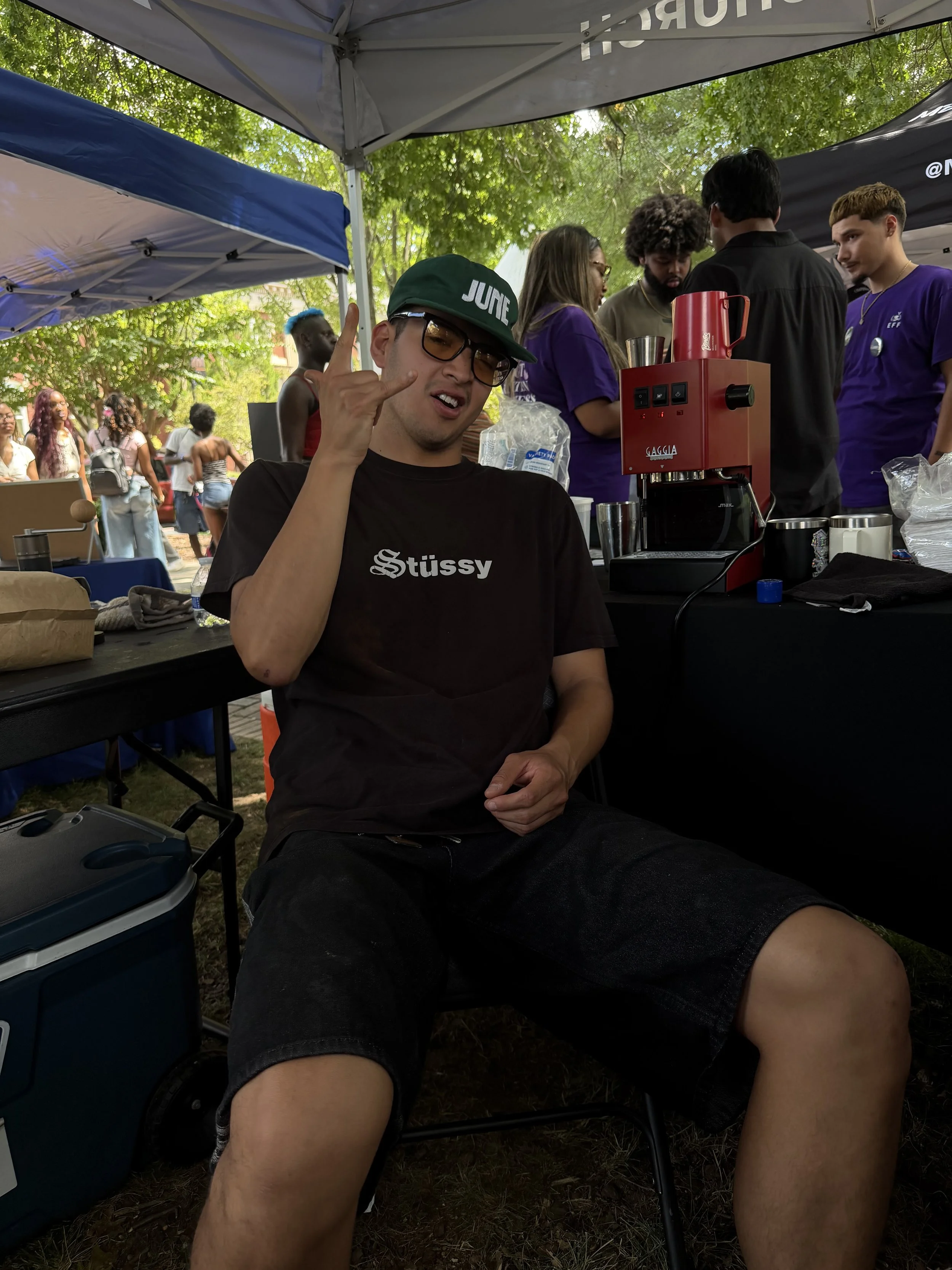 A young man wearing glasses and a black T-shirt with 'Stüssy' written on it, holding up a hand with the index finger and pinky finger extended, sitting under a canopy at an outdoor event. There are trees, other people, and a red coffee machine on a table behind him.