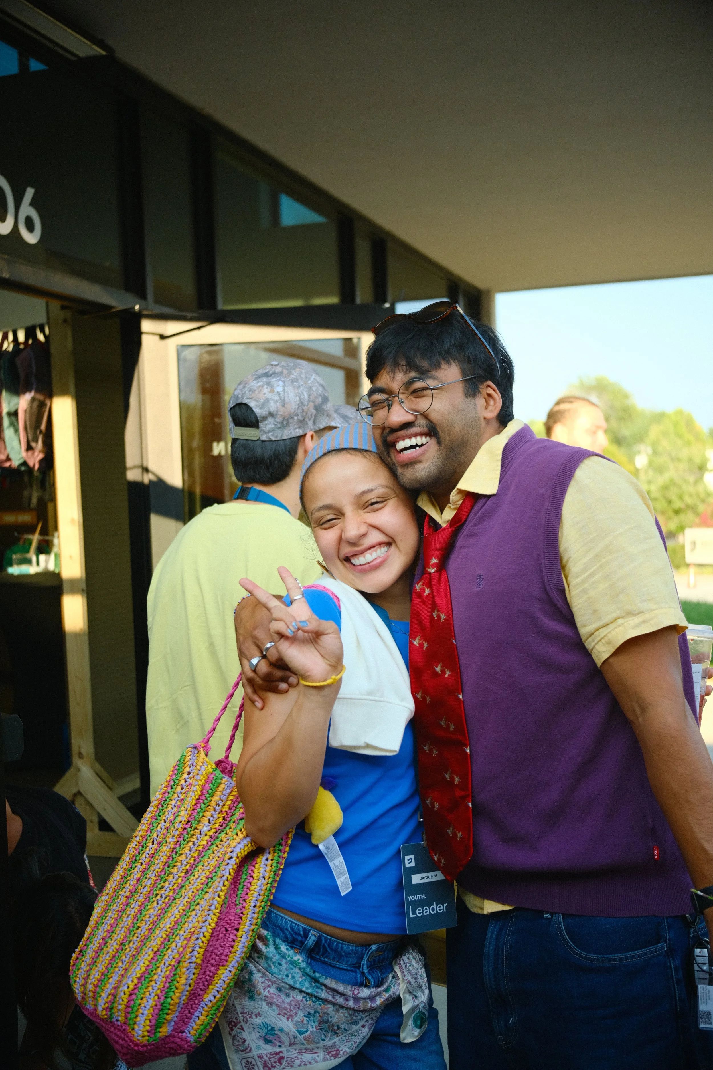 A young girl and a man are smiling and hugging. The girl is wearing a blue and white shirt, multiple bracelets, and a colorful crochet bag. The man is wearing glasses, a yellow shirt, a purple vest, and a red tie. They are outdoors with a building and trees in the background.