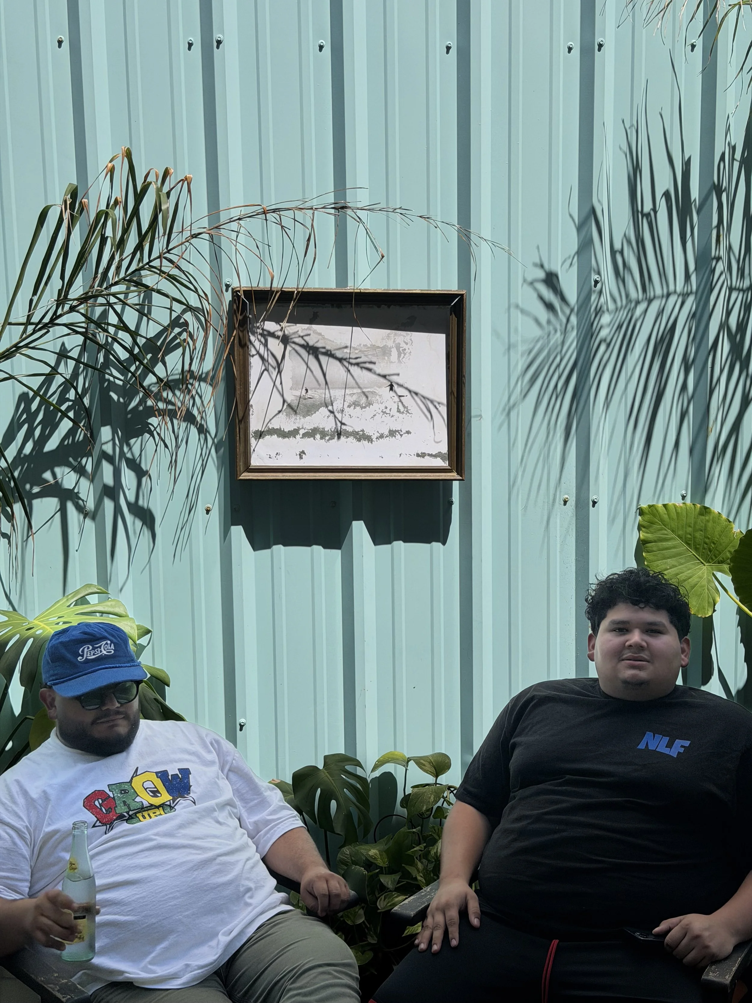 Two men are sitting outdoors against a teal corrugated metal wall, surrounded by green plants. The man on the left is wearing glasses, a blue cap, and a white t-shirt with colorful text, holding a bottle of soda. The man on the right has curly dark hair, a black t-shirt with blue lettering, and is sitting with his arm resting on his leg. The shadows of the plants are cast on the wall behind them.